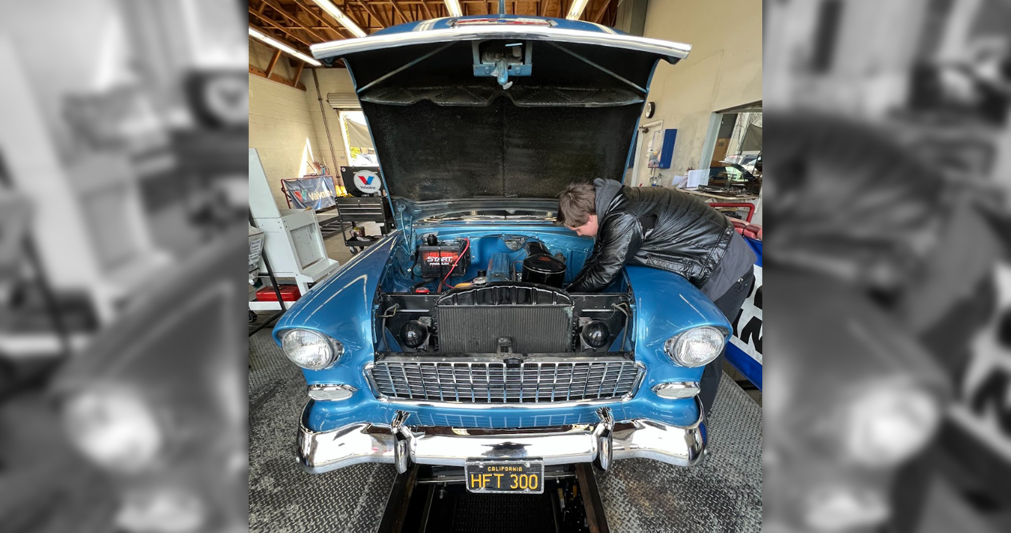 A vintage blue Chevrolet with its hood open being inspected by a technician at an automotive service station.