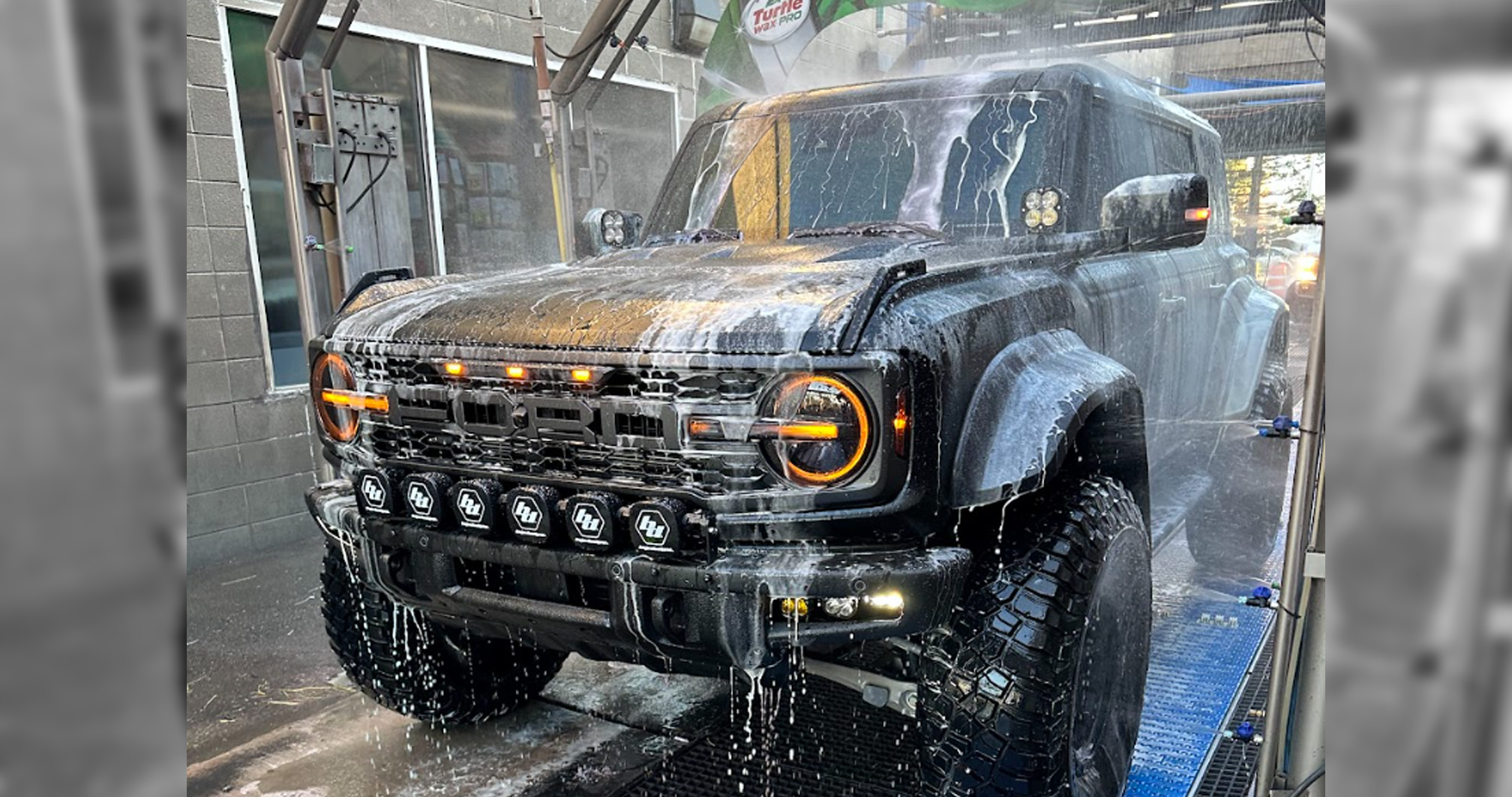 A rugged black Ford Bronco covered in soap and water during a high-pressure car wash, its orange headlights illuminated.