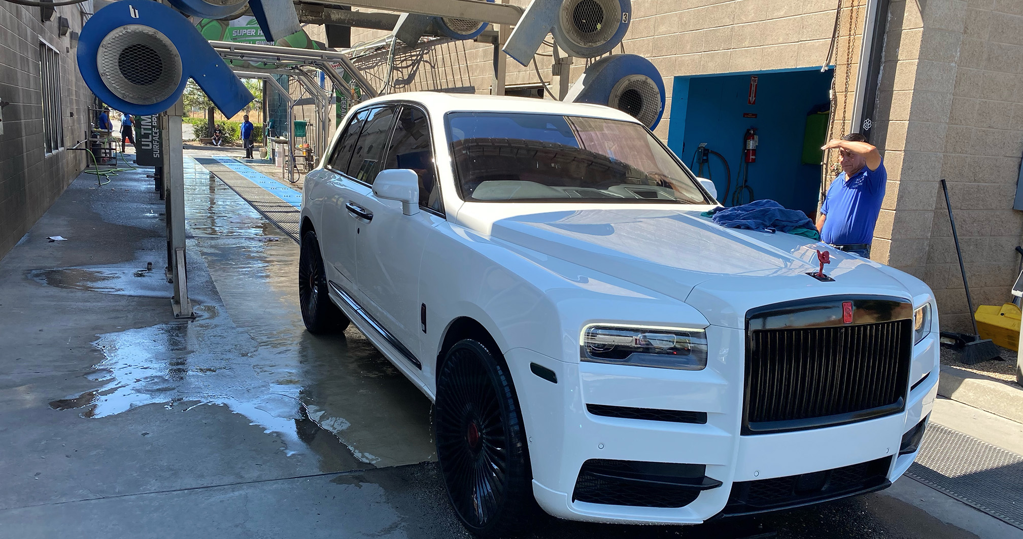 A luxurious white Rolls-Royce Cullinan parked in a car wash drying area, with an attendant wiping it down.